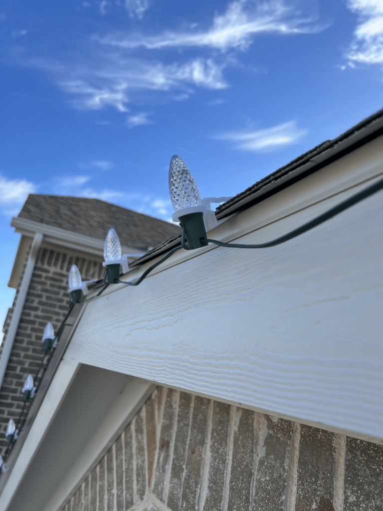 Close-up of a string of clear, LED holiday lights attached to the edge of a house's roofline. The roof has brown shingles and the house is made of light brown brick. The sky is blue with a few scattered clouds, showcasing the brilliance of commercial Christmas lighting perfect for festive decor.