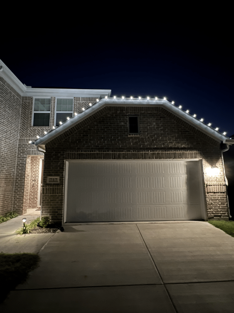 A two-story brick house with a double garage illuminated at night. String lights decorate the roofline above the garage, and the porch and walkway are also lit with wholesale Christmas lights. The garage door is closed, and the driveway is empty.