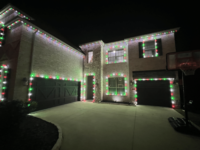 A two-story house is decorated with bulk Christmas lights in red, green, and white, outlining the roof and windows. The lights create a festive holiday atmosphere. There’s a basketball hoop on the right side near the garage. The scene is captured at night.