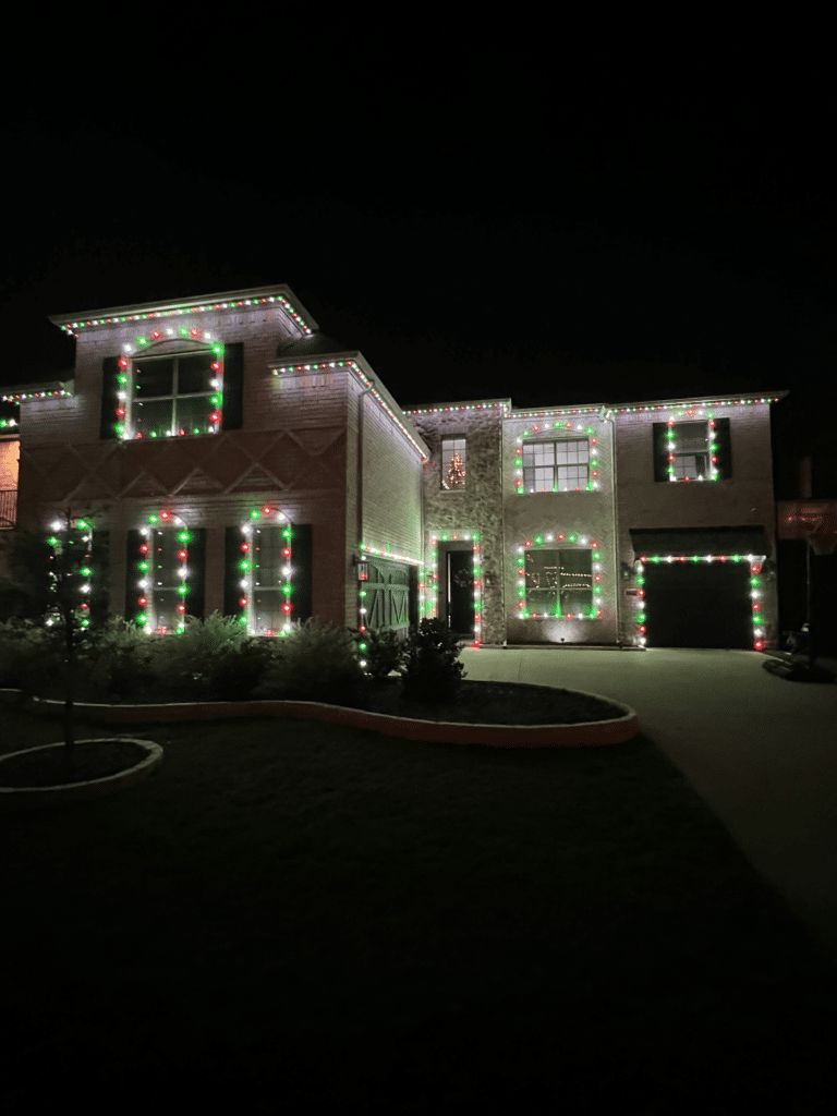 A large two-story house is beautifully decorated with red, white, and green Christmas lights outlining the roof, windows, and garage. The house is surrounded by a well-maintained lawn and shrubs, all set against a dark night sky.